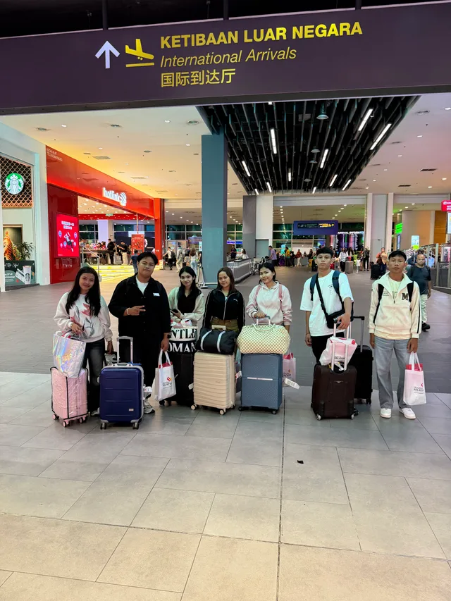 Students with suitcases beneath an international arrivals sign